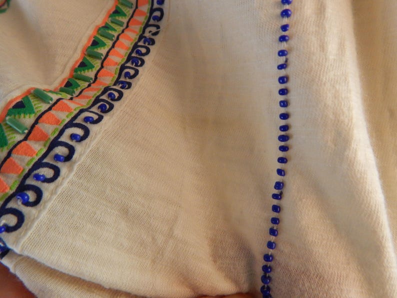 May include: Close-up of a cream-colored fabric with decorative embroidery. The embroidery features a combination of green, orange, and blue colors. A line of blue beads runs vertically along the fabric's edge, adding a touch of elegance.