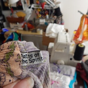 May include: Close-up of a fabric with a floral pattern in shades of peach, green, and purple. A small white tag with the text "Paths of the Spirit" is attached. The background shows a sewing machine and various sewing supplies.