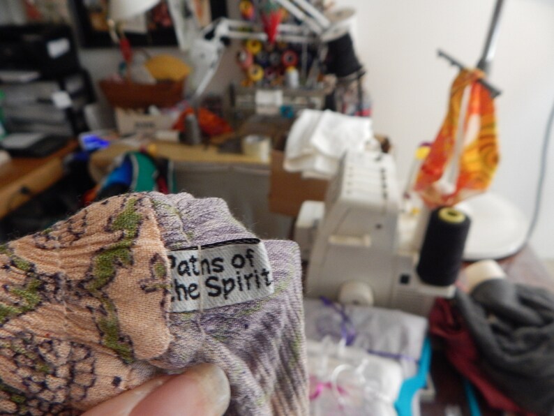 May include: Close-up of a fabric with a floral pattern in shades of peach, green, and purple. A small white tag with the text "Paths of the Spirit" is attached. The background shows a sewing machine and various sewing supplies.