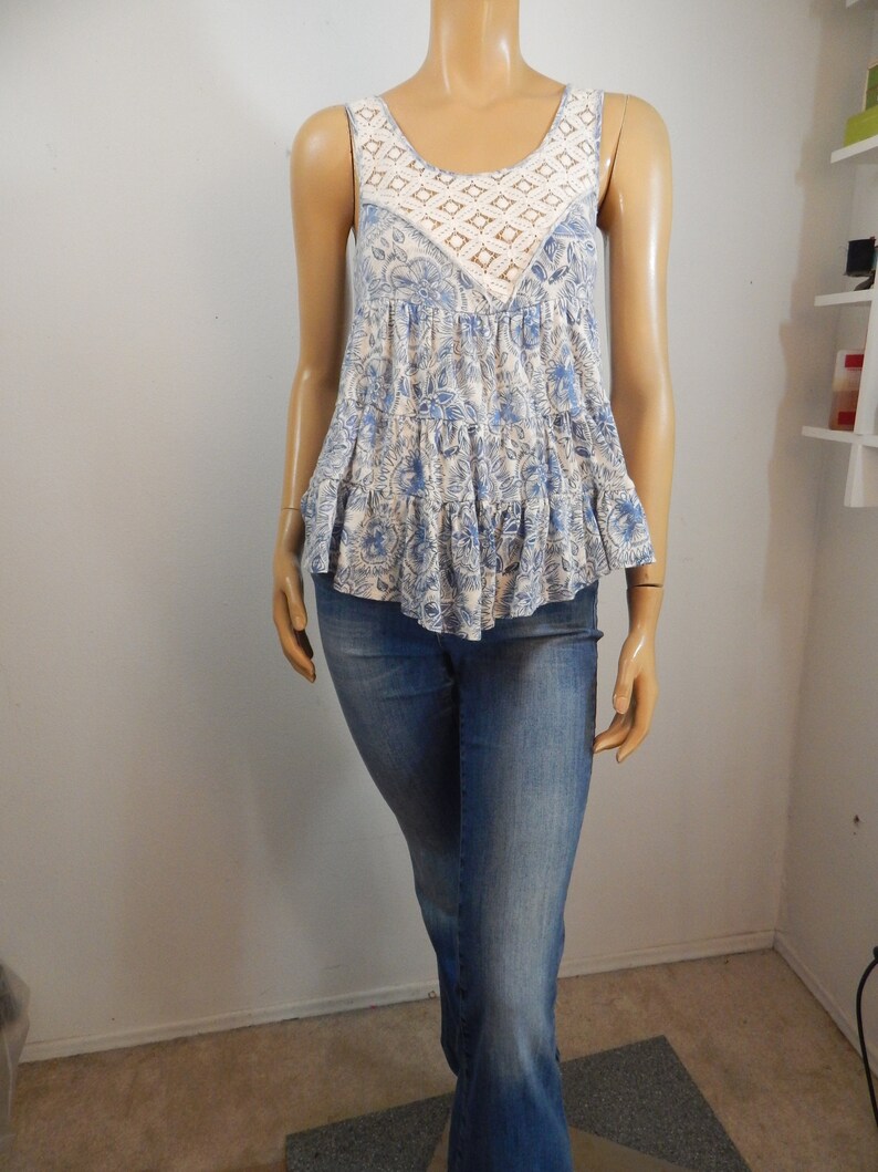 May include: A sleeveless blue and white floral print top with a crochet neckline and a tiered ruffle hem. The top is paired with blue denim jeans.