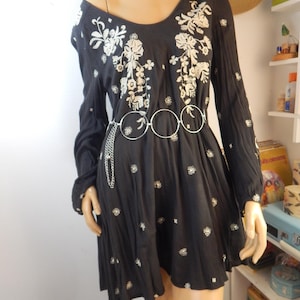 May include: Black long-sleeve dress with white floral embroidery on the bodice and scattered throughout the skirt. The dress features a scoop neckline and a silver-tone belt with three connected circles. The dress is displayed on a mannequin.