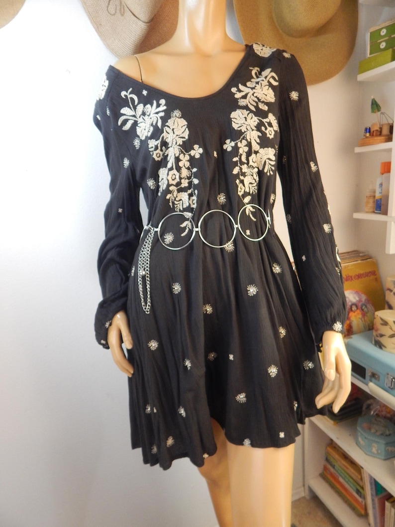 May include: Black long-sleeve dress with white floral embroidery on the bodice and scattered throughout the skirt. The dress features a scoop neckline and a silver-tone belt with three connected circles. The dress is displayed on a mannequin.