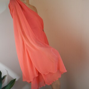 Puede incluir: Un vestido color coral con un solo hombro y un dise&ntilde;o fluido y en capas. El vestido presenta una manga larga y un dobladillo delicado y festoneado. Se puede ver un peque&ntilde;o adorno decorativo en la cintura.