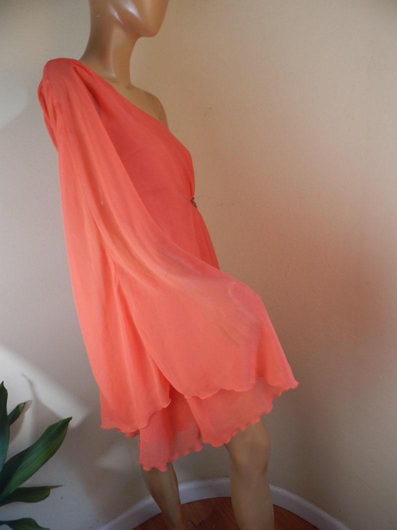 Puede incluir: Un vestido color coral con un solo hombro y un dise&ntilde;o fluido y en capas. El vestido presenta una manga larga y un dobladillo delicado y festoneado. Se puede ver un peque&ntilde;o adorno decorativo en la cintura.
