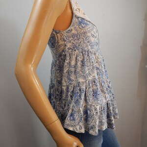 May include: A sleeveless top with a floral pattern in blue and white. The top has a tiered, ruffled design and a lace detail at the neckline. The garment is made of a soft, lightweight fabric.