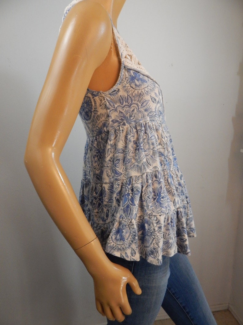 May include: A sleeveless top with a floral pattern in blue and white. The top has a tiered, ruffled design and a lace detail at the neckline. The garment is made of a soft, lightweight fabric.