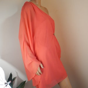 Puede incluir: Vestido coral de un solo hombro con un dise&ntilde;o asim&eacute;trico y fluido. El vestido presenta una manga larga y un detalle en la cintura ce&ntilde;ida. La tela parece ligera y semitransparente, adecuada para climas c&aacute;lidos.