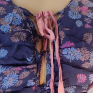 May include: A dark blue floral top with a V-neckline and a pink ribbon tie. The fabric is patterned with blue, pink, and peach-colored flowers. The top has a gathered design.
