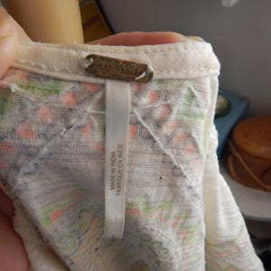 May include: Close-up of a cream-colored top with a floral pattern in orange, green, and purple. The tag reads "MADE IN INDIA FABRIQUE EN INDE". The top is made of a lightweight fabric.