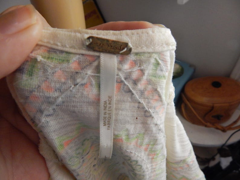 May include: Close-up of a cream-colored top with a floral pattern in orange, green, and purple. The tag reads "MADE IN INDIA FABRIQUE EN INDE". The top is made of a lightweight fabric.