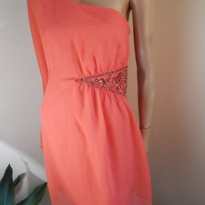 Puede incluir: Vestido color coral con un solo hombro. El vestido tiene una manga larga y un adorno de cuentas en la cintura. El vestido, a la altura de la rodilla, est&aacute; hecho de una tela fluida.