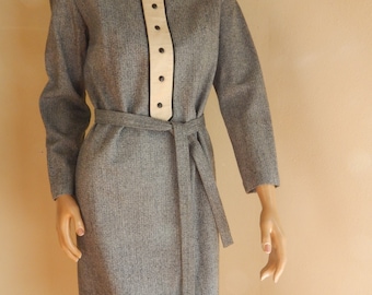 May include: A gray dress with long sleeves and a matching belt. The dress features a cream-colored panel down the front with black buttons and trim. The dress has a round neckline and a knee-length hemline.