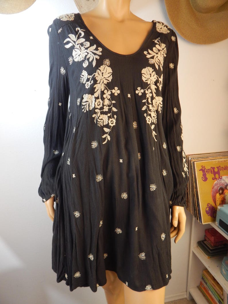 May include: Black long-sleeve dress with a scoop neck and floral embroidery. The dress features white embroidered flowers and small white floral accents. The dress is made of a lightweight fabric and has a loose, flowing fit.