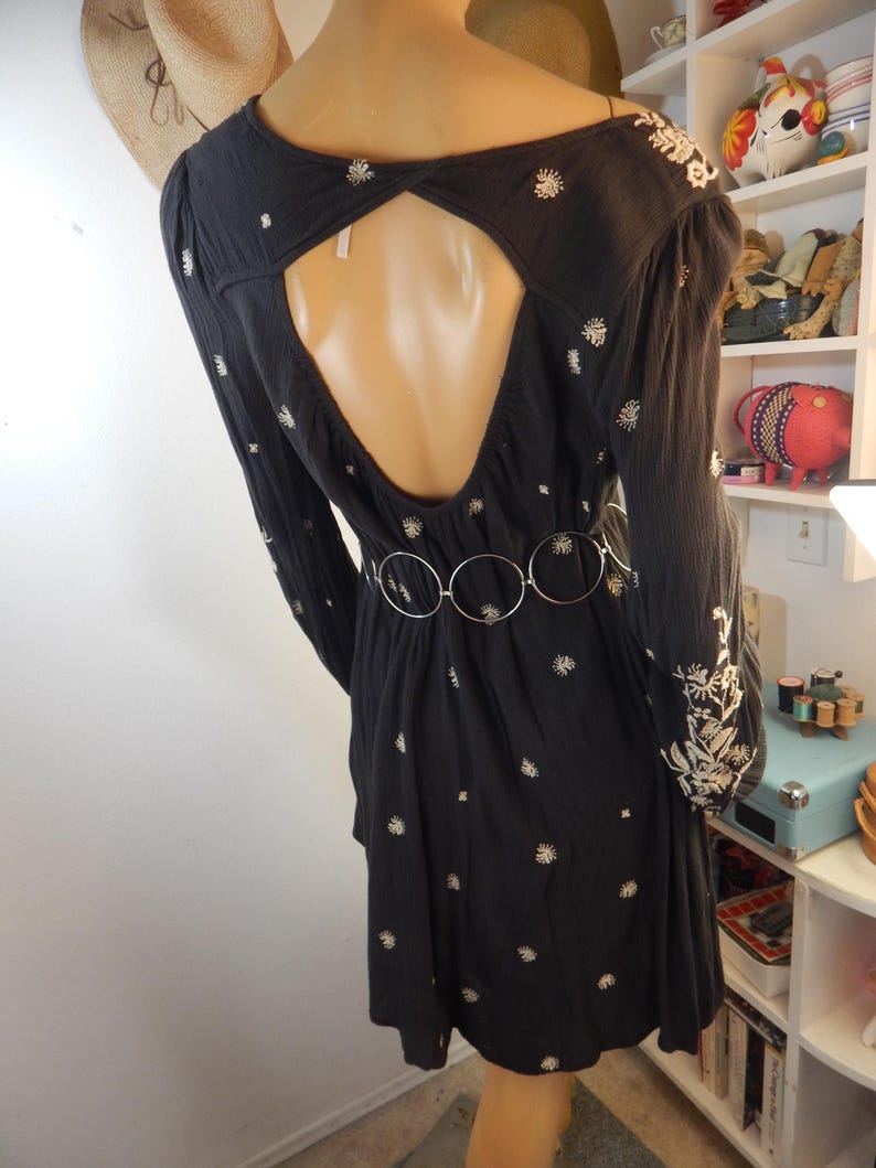May include: Black dress with long sleeves and a keyhole back. The dress features white floral embroidery and a silver belt. The dress is displayed on a mannequin.