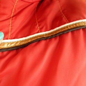 May include: Close-up of a red jacket with gold stitching and a white and gold trim. The jacket has a zipper and is made of a smooth, shiny material. The trim is accented with a thin black line.