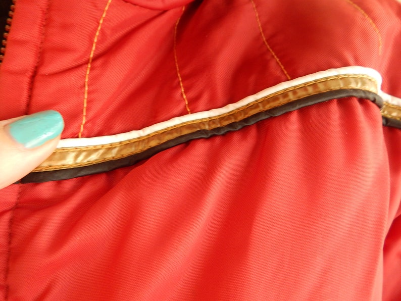 May include: Close-up of a red jacket with gold stitching and a white and gold trim. The jacket has a zipper and is made of a smooth, shiny material. The trim is accented with a thin black line.