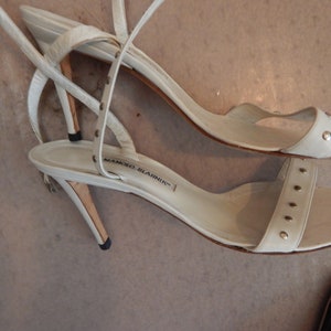 May include: A pair of white leather sandals with thin straps and small silver studs. The sandals have a stiletto heel and the brand name "MANOLO BLAHNIK" is visible on the insole.