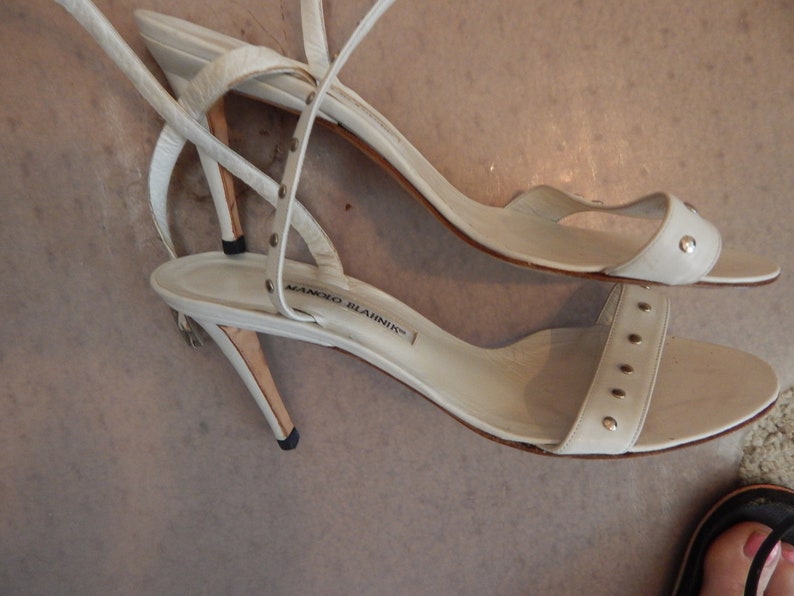 May include: A pair of white leather sandals with thin straps and small silver studs. The sandals have a stiletto heel and the brand name "MANOLO BLAHNIK" is visible on the insole.