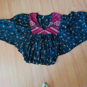 May include: A dark blue top with a floral pattern and wide sleeves. The top features a V-neck with a patterned yoke in shades of red, pink, and purple. The sleeves have pink cuffs. The top is laying on a wooden surface.