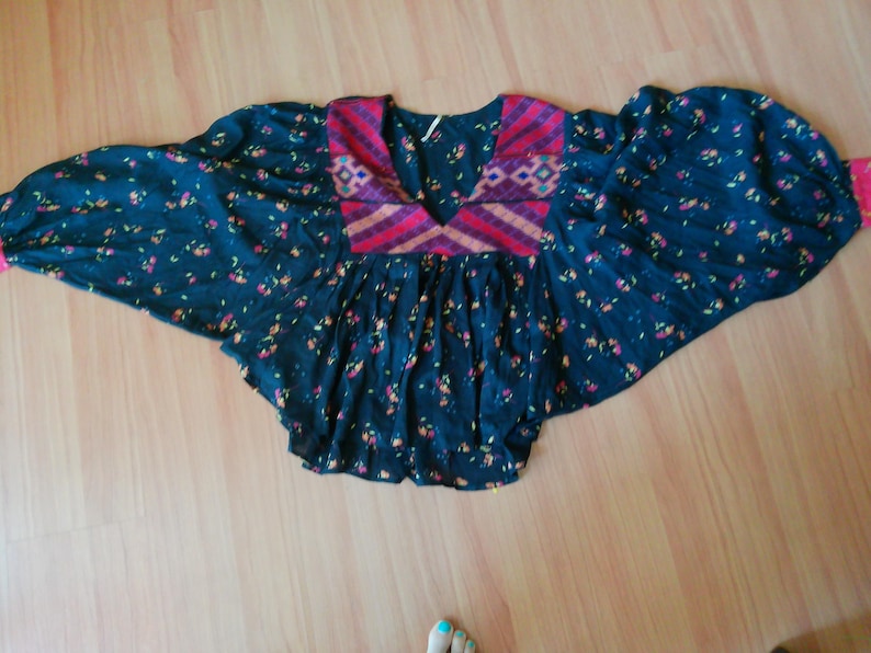 May include: A dark blue top with a floral pattern and wide sleeves. The top features a V-neck with a patterned yoke in shades of red, pink, and purple. The sleeves have pink cuffs. The top is laying on a wooden surface.