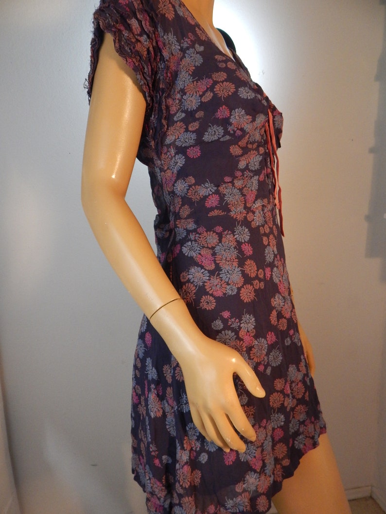 May include: A dark purple dress with a floral pattern in pink, blue, and orange. The dress has short, ruffled sleeves and a V-neckline with a tie. The fabric appears lightweight and flowing, suitable for warm weather.