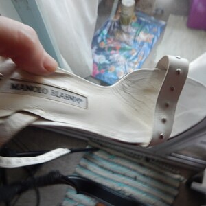 May include: Cream-coloured sandal with a strap adorned with small silver studs. The interior of the shoe features the "MANOLO BLAHNIK" brand name. The shoe is held by a hand.