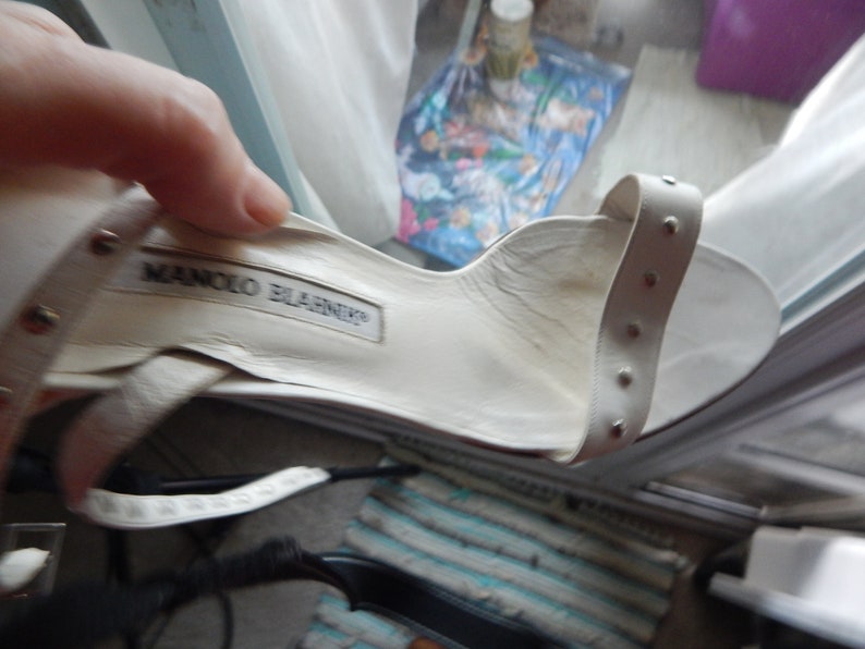 May include: Cream-coloured sandal with a strap adorned with small silver studs. The interior of the shoe features the "MANOLO BLAHNIK" brand name. The shoe is held by a hand.
