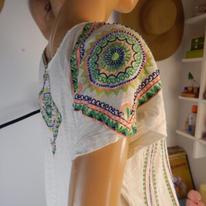 May include: Off-white tunic top with colorful embroidered details. The short-sleeved top features intricate circular and geometric patterns in shades of green, blue, and orange on the shoulders and sleeves. Vertical lines of embroidery run down the front.