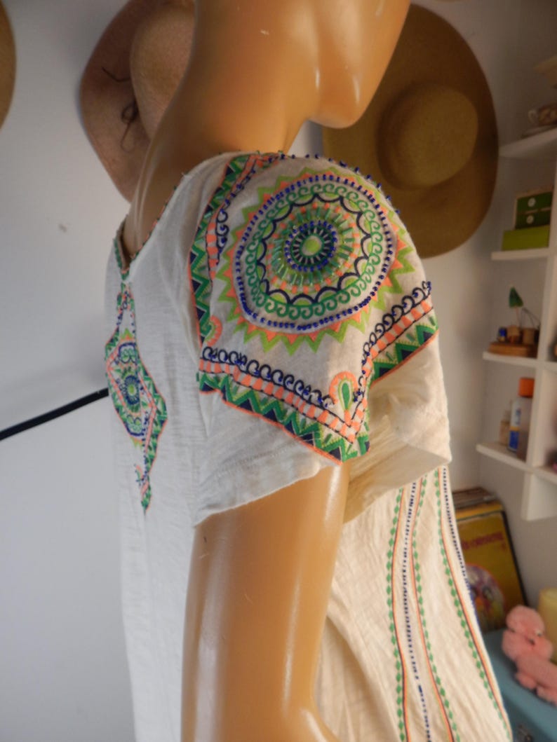 May include: Off-white tunic top with colorful embroidered details. The short-sleeved top features intricate circular and geometric patterns in shades of green, blue, and orange on the shoulders and sleeves. Vertical lines of embroidery run down the front.