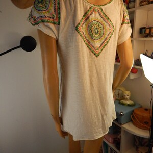 May include: Cream-colored short-sleeve shirt with colorful embroidered details. The shirt features a diamond-shaped design on the back and shoulder accents with green, orange, and blue patterns. The shirt has a scoop neck and a relaxed fit.