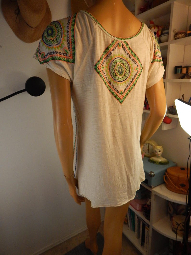 May include: Cream-colored short-sleeve shirt with colorful embroidered details. The shirt features a diamond-shaped design on the back and shoulder accents with green, orange, and blue patterns. The shirt has a scoop neck and a relaxed fit.