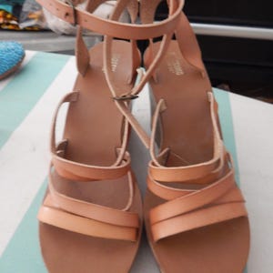 May include: A pair of light brown leather sandals with multiple straps and ankle straps. The sandals have a flat sole and a buckle closure. The sandals are displayed on a striped surface.