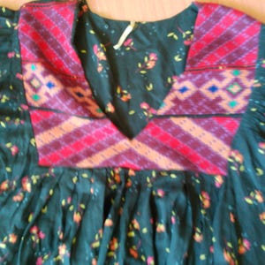 May include: Dark teal dress with floral print and a patterned yoke. The yoke features red, orange, and purple geometric designs. The dress has a round neckline and gathered skirt.