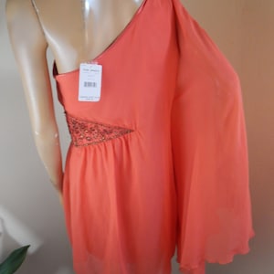 Puede incluir: Vestido coral de un solo hombro con dise&ntilde;o asim&eacute;trico. El vestido tiene una manga larga y un adorno de cuentas en el lateral. Una etiqueta est&aacute; adjunta. El vestido se exhibe en un maniqu&iacute;.