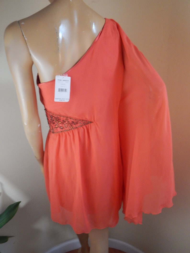 Puede incluir: Vestido coral de un solo hombro con dise&ntilde;o asim&eacute;trico. El vestido tiene una manga larga y un adorno de cuentas en el lateral. Una etiqueta est&aacute; adjunta. El vestido se exhibe en un maniqu&iacute;.