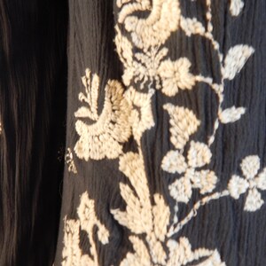 May include: Close-up of a dark navy fabric with intricate off-white floral embroidery. The design features detailed flowers, leaves, and vines, creating a textured and elegant pattern. The fabric has a slightly crinkled appearance.