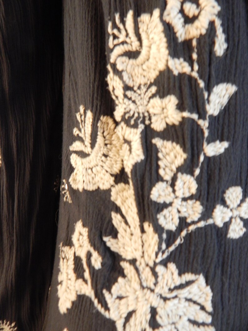 May include: Close-up of a dark navy fabric with intricate off-white floral embroidery. The design features detailed flowers, leaves, and vines, creating a textured and elegant pattern. The fabric has a slightly crinkled appearance.