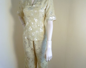May include: A two-piece ensemble featuring a pale yellow top and matching pants. The top has short sleeves and a notched neckline. The fabric is patterned with silver floral designs. The pants are cropped, with a straight leg and a slit at the hem.