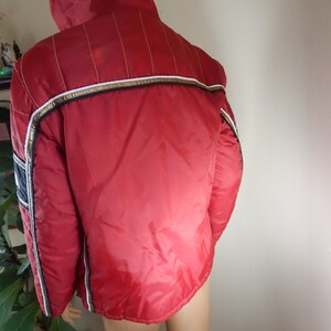 May include: A red vintage jacket with a high collar, white and gold trim, and vertical gold stitching on the shoulders. The jacket has a shiny, quilted appearance and a navy blue stripe on the sleeve.