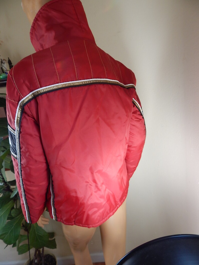 May include: A red vintage jacket with a high collar, white and gold trim, and vertical gold stitching on the shoulders. The jacket has a shiny, quilted appearance and a navy blue stripe on the sleeve.