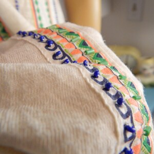 May include: Close-up of a cream-colored garment with decorative trim. The trim features a pattern of orange, green, and blue embroidery and small blue beads. The fabric appears soft and textured.