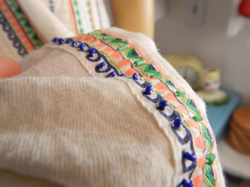 May include: Close-up of a cream-colored garment with decorative trim. The trim features a pattern of orange, green, and blue embroidery and small blue beads. The fabric appears soft and textured.