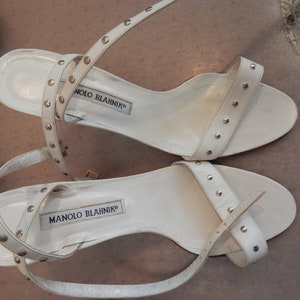 May include: White leather sandals with silver studs. The sandals have thin ankle straps and a small heel. The brand name "MANOLO BLAHNIK" is printed on the insole. The sandals are a stylish footwear choice.