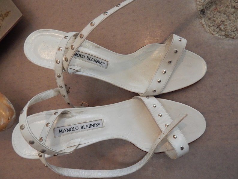 May include: White leather sandals with silver studs. The sandals have thin ankle straps and a small heel. The brand name "MANOLO BLAHNIK" is printed on the insole. The sandals are a stylish footwear choice.