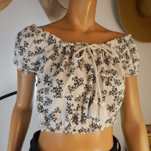 May include: Off-the-shoulder crop top in cream with a black floral pattern. The top features short sleeves and a drawstring neckline with tassel accents. A stylish and feminine piece for casual wear.