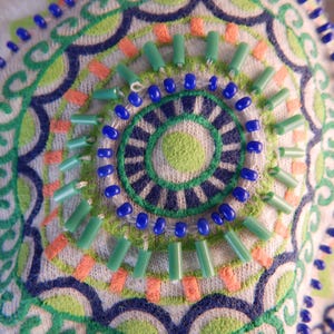 May include: Close-up of a white fabric with an embroidered design. The design features a circular pattern with blue, green, and orange accents. Small blue beads and green tube beads are sewn onto the fabric, adding texture.