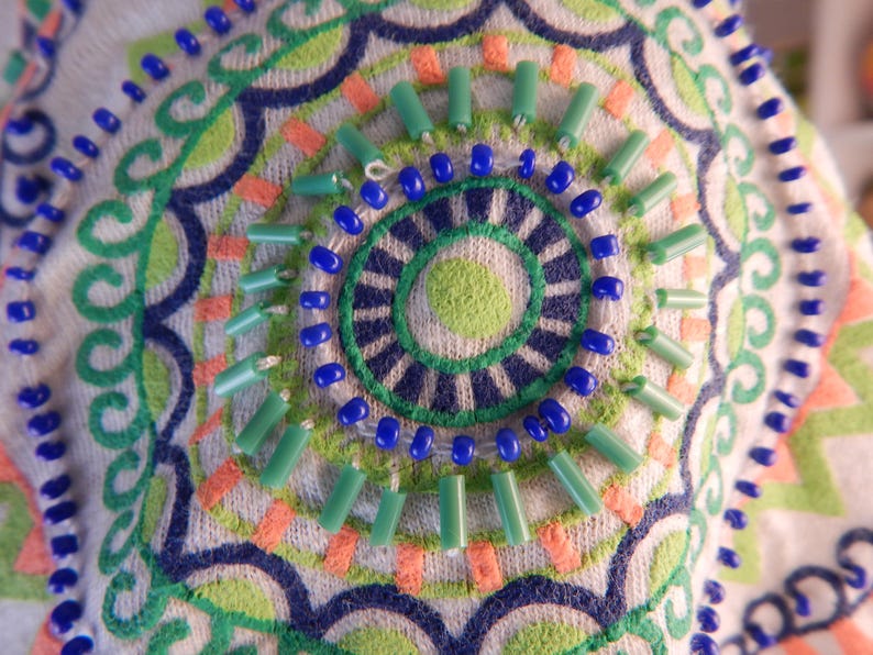 May include: Close-up of a white fabric with an embroidered design. The design features a circular pattern with blue, green, and orange accents. Small blue beads and green tube beads are sewn onto the fabric, adding texture.