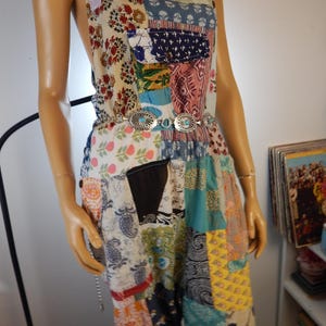 May include: A patchwork jumpsuit in various colors and patterns, including floral, paisley, and geometric designs. The jumpsuit features a silver-tone belt with turquoise accents. The straps are light pink, and the garment appears to be made of multiple fabric pieces.