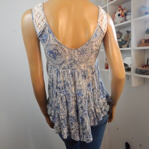 May include: A sleeveless top with a blue and white floral pattern and a ruffled hem. The top features white lace detailing on the shoulders and a low scoop back. The mannequin is wearing blue jeans.
