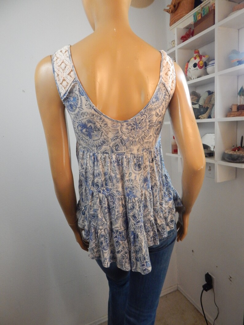 May include: A sleeveless top with a blue and white floral pattern and a ruffled hem. The top features white lace detailing on the shoulders and a low scoop back. The mannequin is wearing blue jeans.